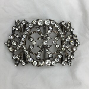 May include: A silver and crystal buckle with a decorative floral design. The buckle is set with clear crystals and has a delicate, intricate design.