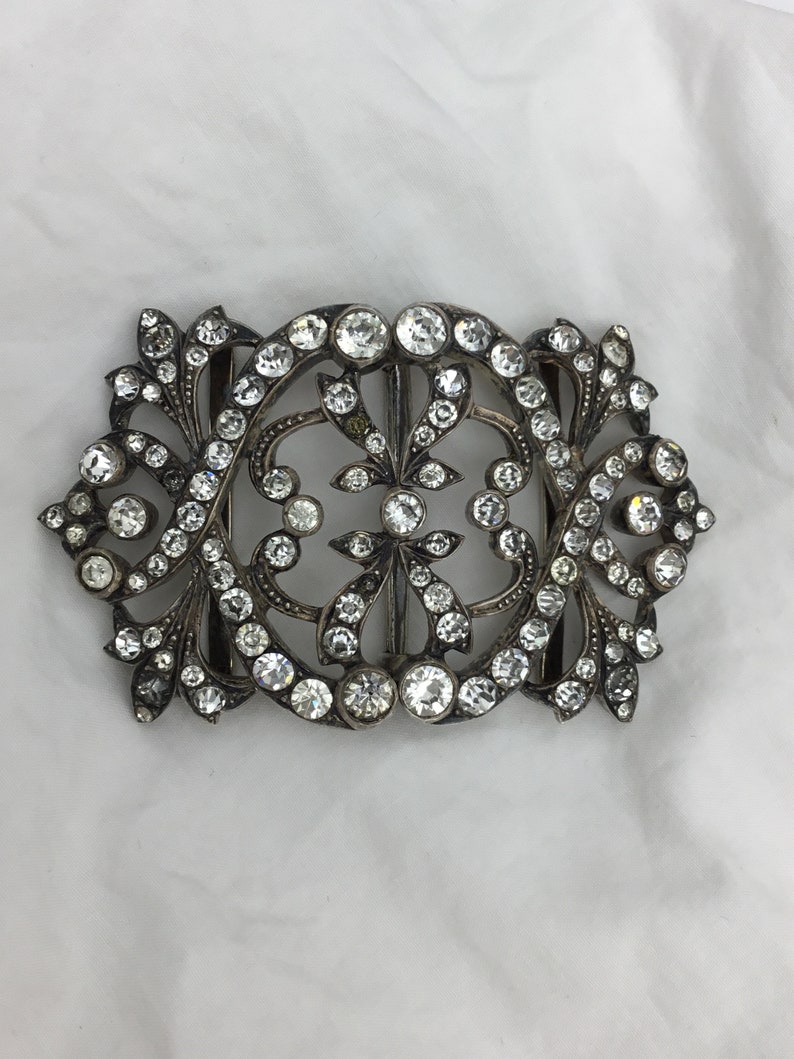 May include: A silver and crystal buckle with a decorative floral design. The buckle is set with clear crystals and has a delicate, intricate design.