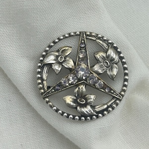 Op de afbeelding: Een zilverkleurige broche met een stervormig ontwerp en drie gestileerde bloemen. De broche is versierd met heldere strasssteentjes.