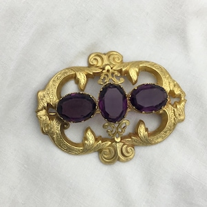 May include: A gold-coloured brooch featuring three oval, deep purple gemstones. The brooch has an ornate, filigree design with floral and scrollwork details. The gemstones are set in a decorative frame.