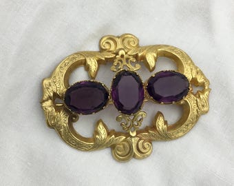 May include: A gold-toned brooch with three oval, deep purple gemstones. The brooch has an ornate, filigree design with floral and scrollwork details. The gemstones are set in a decorative frame.