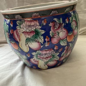 Peut inclure: Un cache-pot en céramique rond avec une base bleu profond, orné d'un motif coloré de fruits roses et rouges, de feuilles vertes et de fleurs blanches. Le bord supérieur est noir avec une bordure blanche et un motif floral blanc et vert.