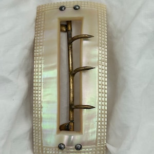 Puede incluir: Una hebilla de cintur&oacute;n de n&aacute;car blanco con un gancho de lat&oacute;n y cuatro puntas. La hebilla tiene una superficie texturizada y est&aacute; decorada con tachuelas plateadas.