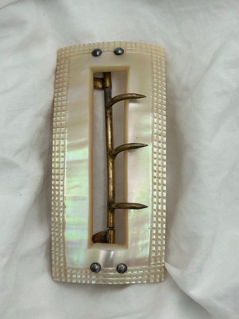 Puede incluir: Una hebilla de cintur&oacute;n de n&aacute;car blanco con un gancho de lat&oacute;n y cuatro puntas. La hebilla tiene una superficie texturizada y est&aacute; decorada con tachuelas plateadas.