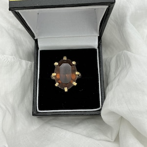 Puede incluir: Un anillo dorado con una gran piedra preciosa ovalada de color marrón. El anillo está engastado en una banda dorada con puntas decorativas. El anillo se muestra en una caja de joyería negra con interior blanco.