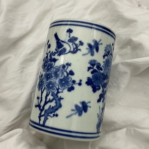May include: A cylindrical white ceramic vase with blue floral and bird designs. The vase has a blue band at the top and bottom. The detailed illustrations include flowers, butterflies, and a bird perched on a branch.