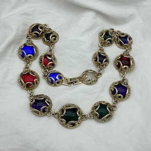 May include: A gold-tone metal chain necklace featuring a series of spherical links. Each link contains a translucent glass bead in shades of red, blue, green, and purple, encased in an intricate, swirling filigree metal cage design.