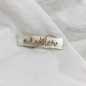 May include: A white mother's day brooch with the word "Mother" written in gold wire.