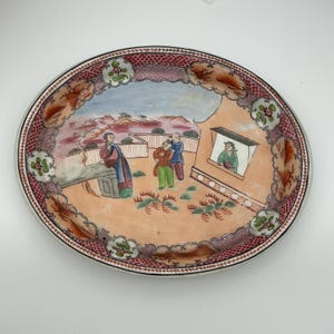 May include: An antique, oval-shaped porcelain plate with a hand-painted scene of figures in traditional attire. The plate features a red and white patterned border and a peach-colored background.