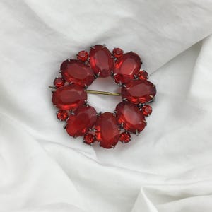 Pode incluir: Broche vintage com um design circular de grandes pedras preciosas vermelhas ovais. Pedras vermelhas menores são intercaladas entre as maiores. O broche tem uma moldura de metal prateado e um alfinete dourado.