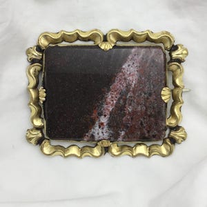 May include: A gold-toned brooch with a rectangular, brown, white, and red agate stone set in the center. The brooch has a decorative, ornate frame.