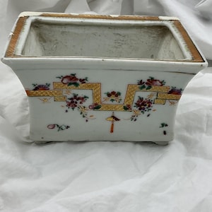 May include: Rectangular white ceramic planter with a gold rim and decorative floral and geometric patterns. The planter features a slightly flared shape and small feet. The floral design includes red, purple, and yellow accents.