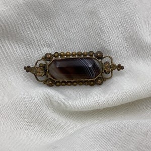 Piccola spilla antica di epoca georgiana-vittoriana in agata marrone fasciata e ottone decorato. Dimensioni: 4,6 cm x 1,5 cm nel punto più largo. Dettagli a foglia. Decorata.