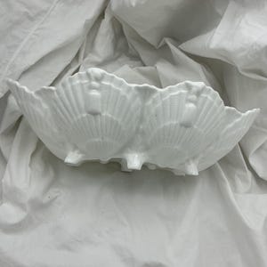 May include: A white ceramic bowl shaped like a series of connected scallop shells. The exterior features a textured, ribbed pattern radiating from the base of each shell section, with small feet supporting the structure on the bottom surface.