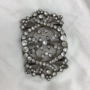 May include: A silver and crystal ornate buckle with a detailed design of intertwined swirls and leaves. The buckle is covered in clear rhinestones.