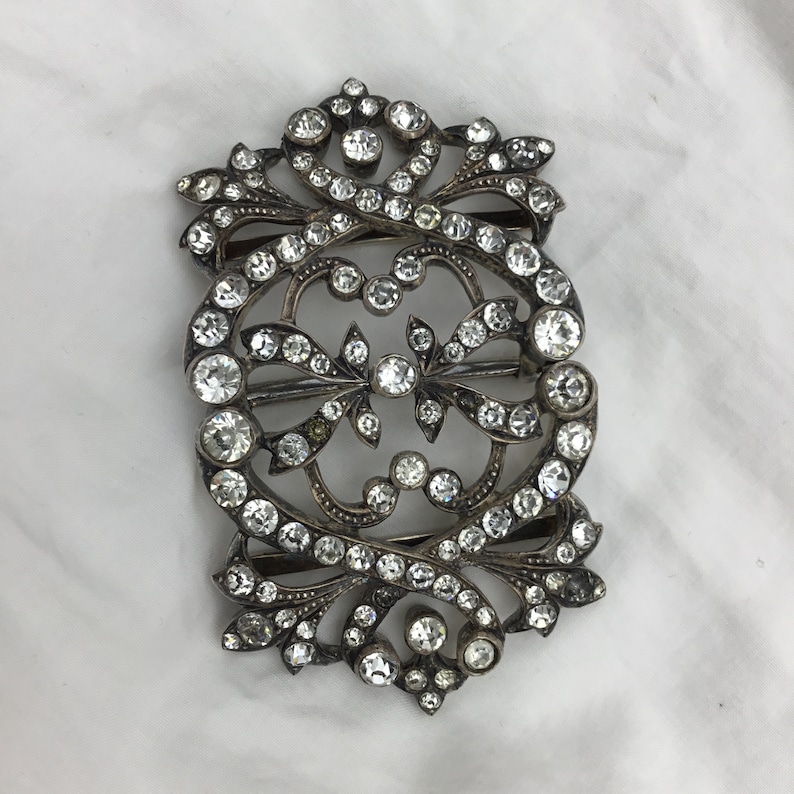May include: A silver and crystal ornate buckle with a detailed design of intertwined swirls and leaves. The buckle is covered in clear rhinestones.