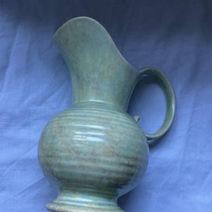 May include: A ceramic pitcher with a rounded body and a flared spout. The surface features a mottled sage green glaze with subtle horizontal ribbed textures around the midsection and a smooth, curved handle extending from the side to the neck.