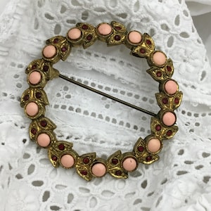 Op de afbeelding: Vintage goudkleurige broche met een cirkelvormig ontwerp. De broche heeft een kransachtig patroon met afwisselend perzikgekleurde stenen en kleine rode accenten. Aan de achterkant is een speld bevestigd om de broche aan kleding of accessoires te bevestigen.
