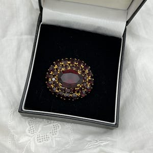 Peut inclure: Une broche de forme ovale avec une grande pierre précieuse centrale rouge foncé entourée de plusieurs petites pierres rouges serties dans une monture en métal doré. La broche est présentée dans un écrin à bijoux doublé de velours noir.