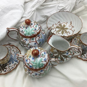 May include: A vintage tea set with a floral design. The set includes a teapot, creamer, sugar bowl, two cups, and two saucers. The pieces are white with gold accents and feature a delicate floral pattern in shades of blue, green, and red.
