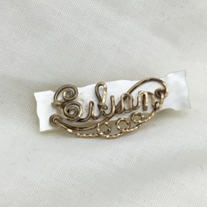 May include: A gold-colored wire name pin that reads "Eulian" in a cursive font. The pin is attached to a small, irregular piece of white material. The pin has decorative swirls and a twisted wire design.