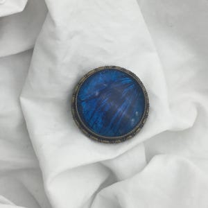 May include: A circular brooch featuring a vibrant blue, iridescent centre, possibly a butterfly wing, set within an antique-style, brass-toned metal frame. The brooch sits on a white fabric, showcasing its intricate design. A unique piece of jewellery.