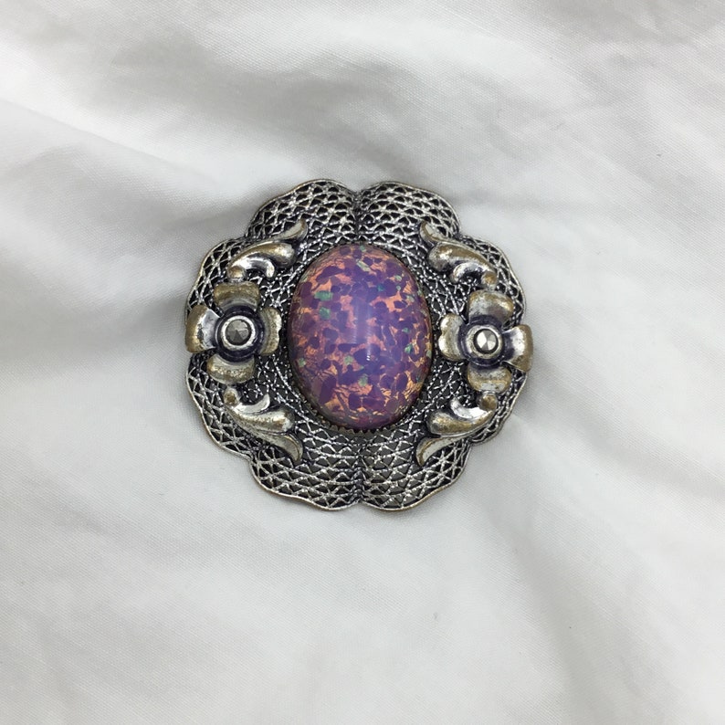 Puede incluir: Un broche de plata con una piedra ovalada p&uacute;rpura iridiscente engastada en un dise&ntilde;o de filigrana.