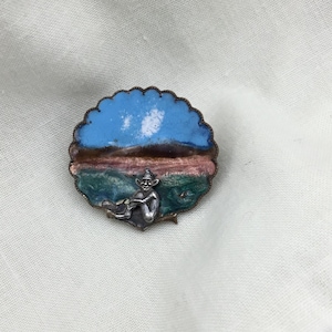 Peut inclure: Une broche en argent et émail représentant une figure fantaisiste assise sur un paysage avec un ciel bleu, des collines vertes et un horizon brun.