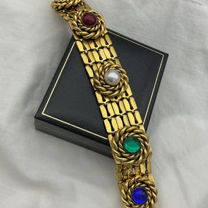 Op de afbeelding: Goudkleurige armband met rechthoekige schakels. De armband is versierd met decoratieve rozetten, elk met een anders gekleurde steen: zwart, rood, wit, groen en blauw. De armband ligt op een zwarte doos.