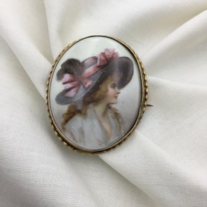 Pu&ograve; includere: Broche ovale in cammeo di porcellana con una montatura dorata. Il cammeo presenta un ritratto dipinto di una donna con un cappello rosa e nero.
