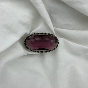 Puede incluir: Broche ovalado con una gema facetada de color púrpura intenso engastada en un marco de metal plateado. El marco tiene un borde decorativo y texturizado. El broche se muestra sobre un fondo de tela blanca texturizada.