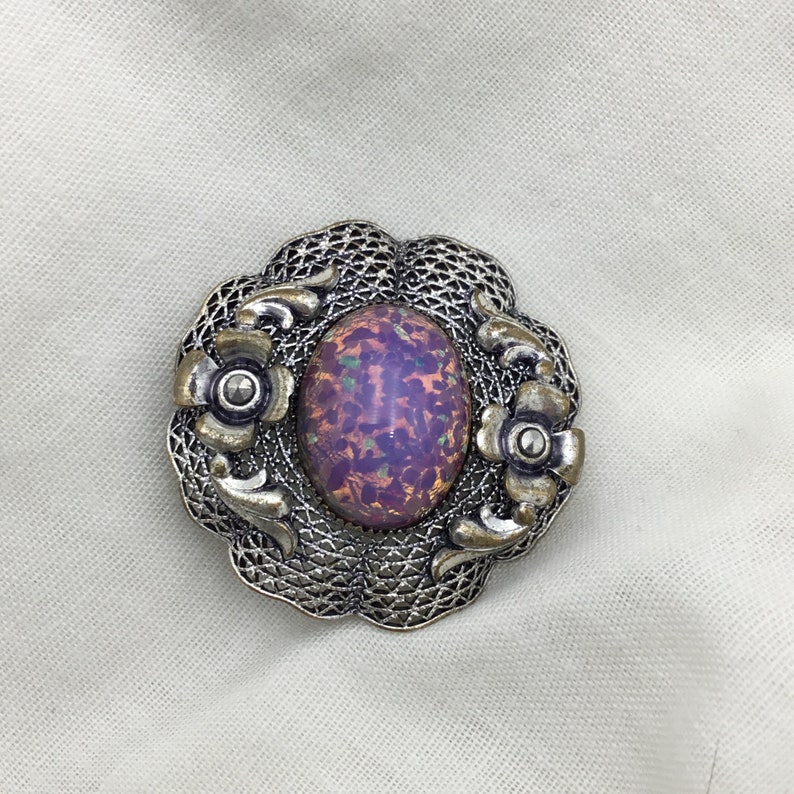 Puede incluir: Un broche de plata con una piedra de &oacute;palo p&uacute;rpura iridiscente de forma ovalada rodeada por un delicado dise&ntilde;o de filigrana. El broche presenta dos peque&ntilde;os detalles de flores a cada lado de la piedra.