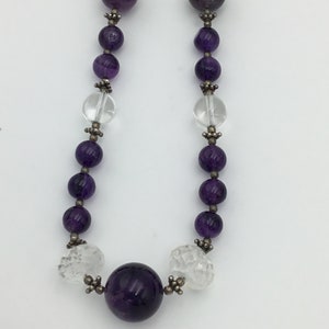 May include: A purple amethyst and clear crystal beaded necklace with silver accents. The necklace features a large, round amethyst bead in the centre.