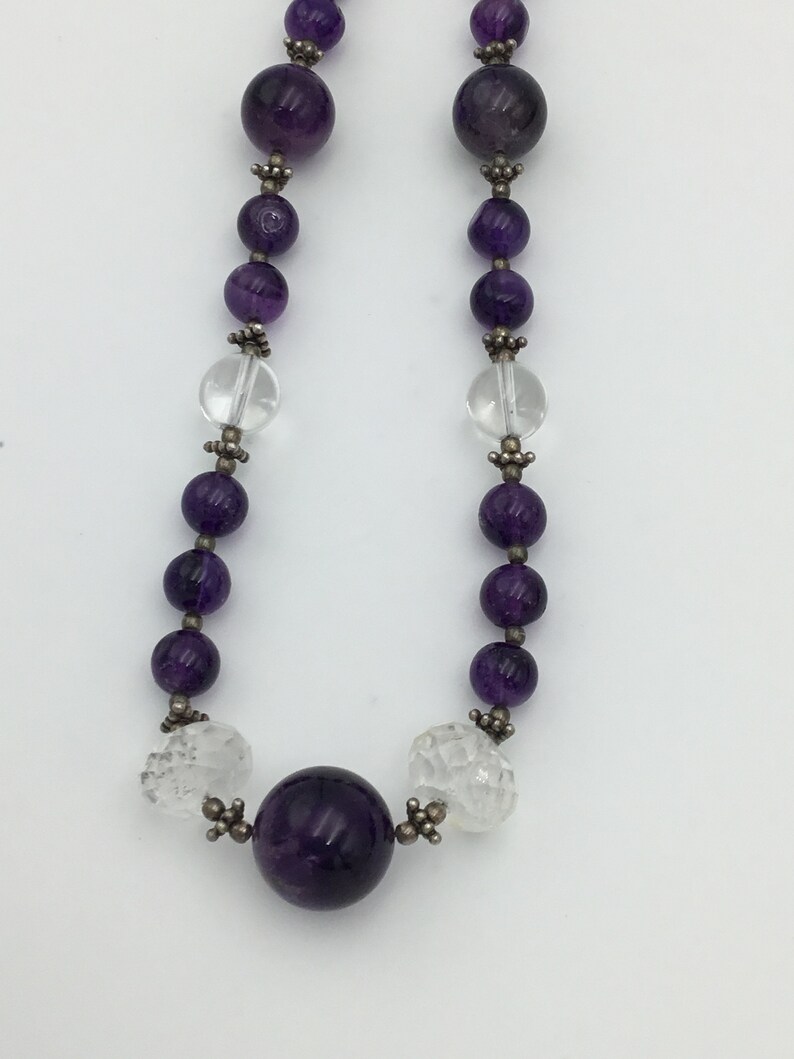 May include: A purple amethyst and clear crystal beaded necklace with silver accents. The necklace features a large, round amethyst bead in the centre.
