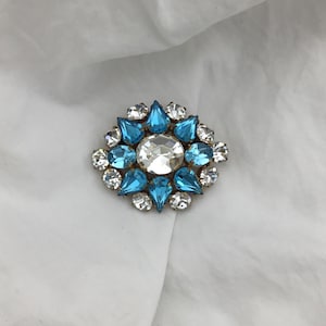 Puede incluir: Broche vintage con diseño floral. El broche presenta una gran piedra central redonda y transparente, rodeada de piedras azul claro en forma de lágrima y piedras más pequeñas y transparentes. El engaste es dorado.