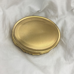 Può includere: Un compatto ovale color oro con un design decorativo testurizzato. Il compatto ha un coperchio incernierato e un piccolo chiavistello.