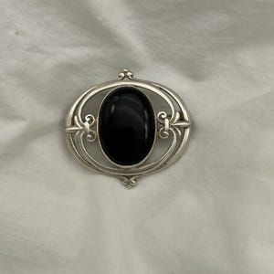 May include: Oval silver brooch with a large, polished black stone centre. The brooch features an ornate, openwork design with decorative flourishes. The setting is a bright, reflective silver, and the stone is a deep, glossy black.