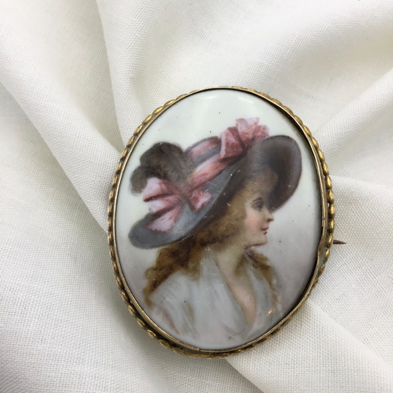 Pu&ograve; includere: Un'antica spilla in porcellana a cammeo raffigurante una donna con un cappello a tesa larga con un nastro rosa. Il cammeo &egrave; incastonato in una cornice in metallo dorato.