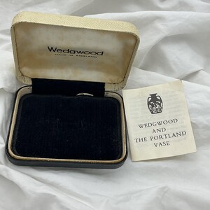 May include: A rectangular hinged presentation box with a cream-coloured textured exterior and a black velvet interior lining. The lid interior features the text Wedgwood Made in England. An accompanying paper pamphlet reads Wedgwood and the Portland Vase.