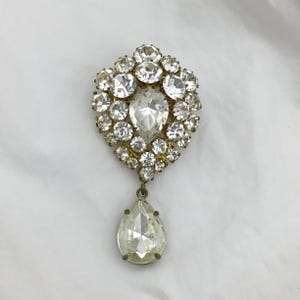 Puede incluir: Broche vintage con un grupo de brillantes redondos y transparentes que rodean un gran brillante en forma de pera. El broche tiene un engaste de metal dorado y un colgante de brillante en forma de pera. Una joya clásica.