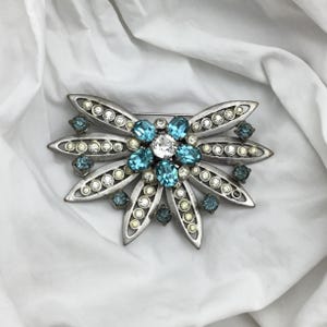 May include: A vintage silver-toned brooch in a floral design. The brooch features a central cluster of blue and white gemstones, surrounded by elongated petals set with clear and blue crystals. A classic accessory.
