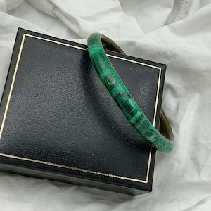 May include: A green malachite bangle bracelet is displayed on a black jewellery box with gold trim. The bracelet has a swirling pattern of dark and light green shades. The box is closed and rests on white fabric.