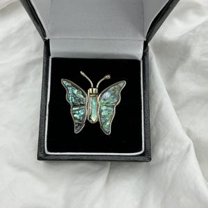 May include: A butterfly-shaped brooch with iridescent wings in a black jewelry box. The wings display shades of green, blue, and silver. The brooch has a silver-colored body and antennae. The box has a black exterior and a white interior.