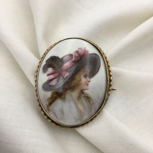 Pu&ograve; includere: Broche ovale in porcellana con cornice dorata. La broche presenta un ritratto dipinto di una donna con un grande cappello a tesa larga con un nastro rosa.