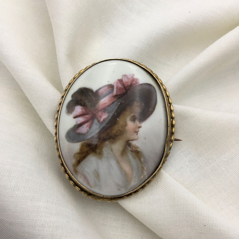 Pu&ograve; includere: Broche ovale in porcellana con cornice dorata. La broche presenta un ritratto dipinto di una donna con un grande cappello a tesa larga con un nastro rosa.
