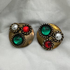 Puede incluir: Par de pendientes vintage de clip. Cada pendiente presenta una base circular dorada con tres elementos decorativos: una gema verde, una gema roja y una perla de imitación, todo ello rodeado por un diseño de cuerda trenzada.