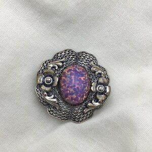Puede incluir: Un broche plateado con una piedra de vidrio p&uacute;rpura iridiscente de forma ovalada engastada en un dise&ntilde;o de filigrana con acentos florales.