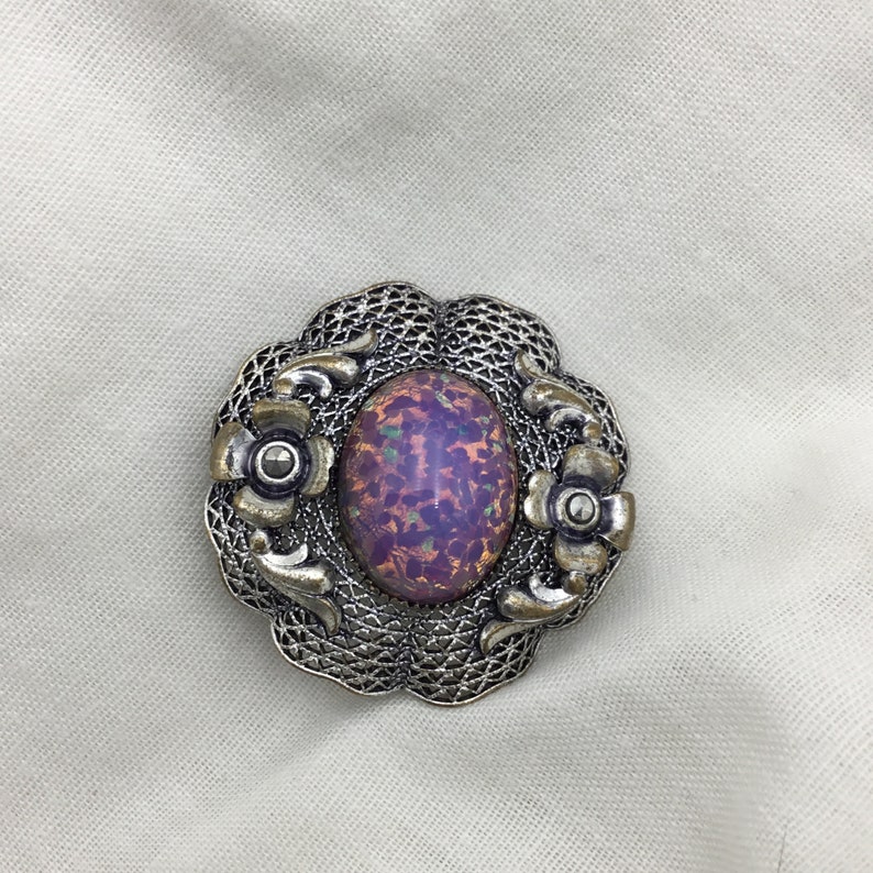 Puede incluir: Un broche plateado con una piedra de vidrio p&uacute;rpura iridiscente de forma ovalada engastada en un dise&ntilde;o de filigrana con acentos florales.