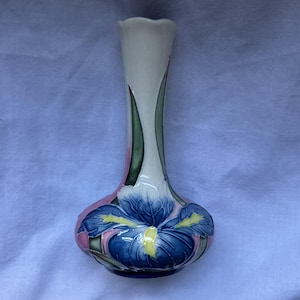 Könnte beinhalten: Eine Keramikvase mit schlankem Hals und bauchigem Fuß, verziert mit einem handgemalten Motiv einer großen blauen Irisblüte mit gelben Zentren. Der Hintergrund der Vase zeigt sanfte Rosa- und Grüntöne, mit einer gewellten Öffnung am Rand.
