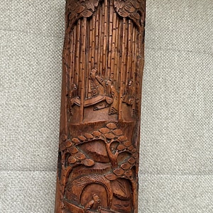 May include: A cylindrical brown wooden brush holder featuring intricate relief carvings of a bamboo forest, figures in traditional robes, a small boat on water, and stylized trees. The surface displays detailed textures and deep wood grain.